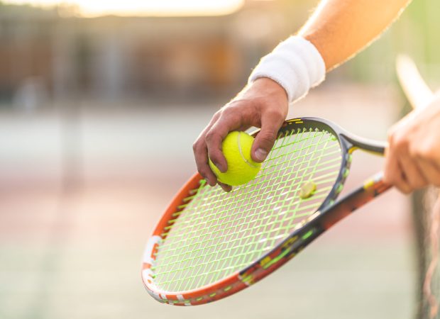 Young Man Playing Tennis Outdoors. Close Up of Tennis Player Hands Holding Racket with Ball. Professional Tennis Player Hitting the Ball. Sport Concept.